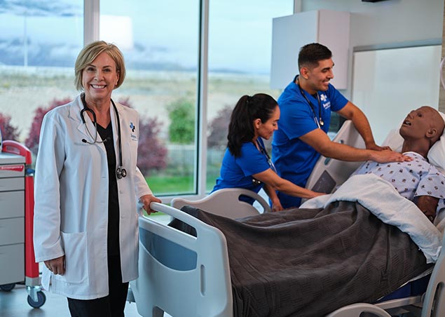 Joyce University faculty in white lab coat smiling while two nursing students in blue scrubs work in the Joyce Johnson simulation center