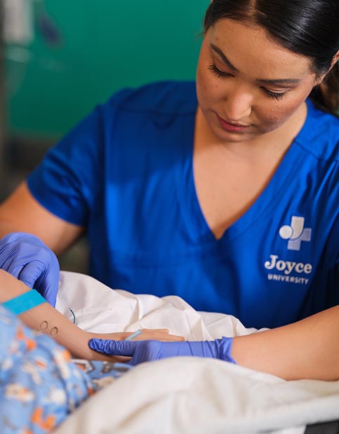 Nursing student in blue Joyce University scrubs inserting IV on high-fidelity mannequin in the simulation lab