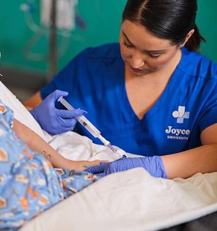 Joyce University student in blue scrubs practicing flushing an IV on pediatric high fidelity mannequin