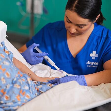 Female nursing student in blue Joyce University scrubs flushing an IV on a pediatric mannequin in the Joyce Johnson simulation lab