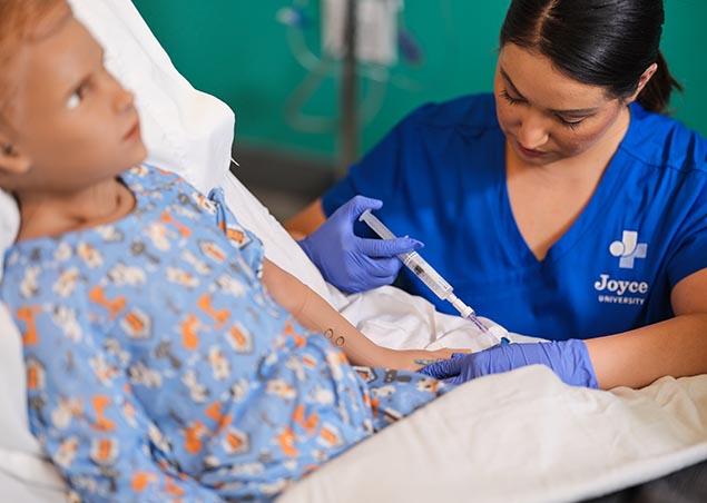 Female nursing student in blue Joyce University scrubs flushing an IV on a pediatric mannequin in the Joyce Johnson simulation lab