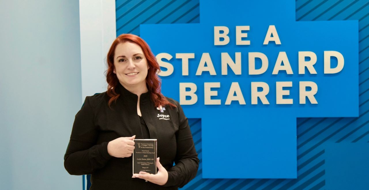 Joyce university faculty standing in front of the Standard Bearer wall holding AACN award