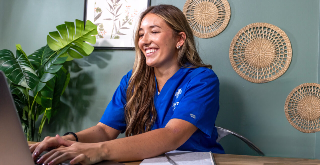Woman in blue joyce university scrubs sitting at a desk in front of a computer