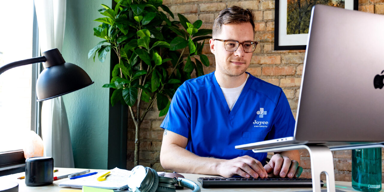 joyce university student in blue nursing scrubs sitting in home office learning remotely at a laptop