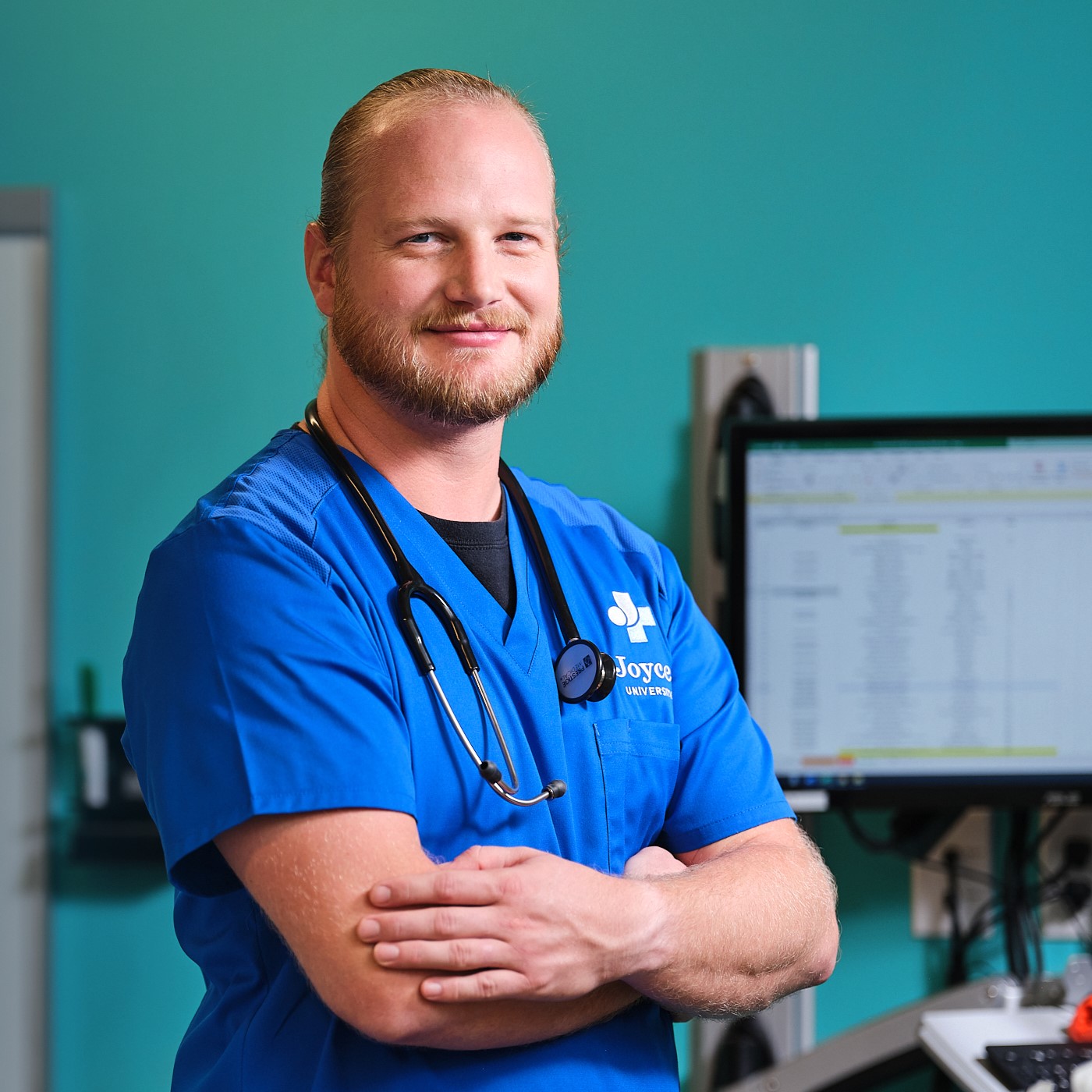 Joyce nursing student in blue scrubs standing in sim room with arms crossed