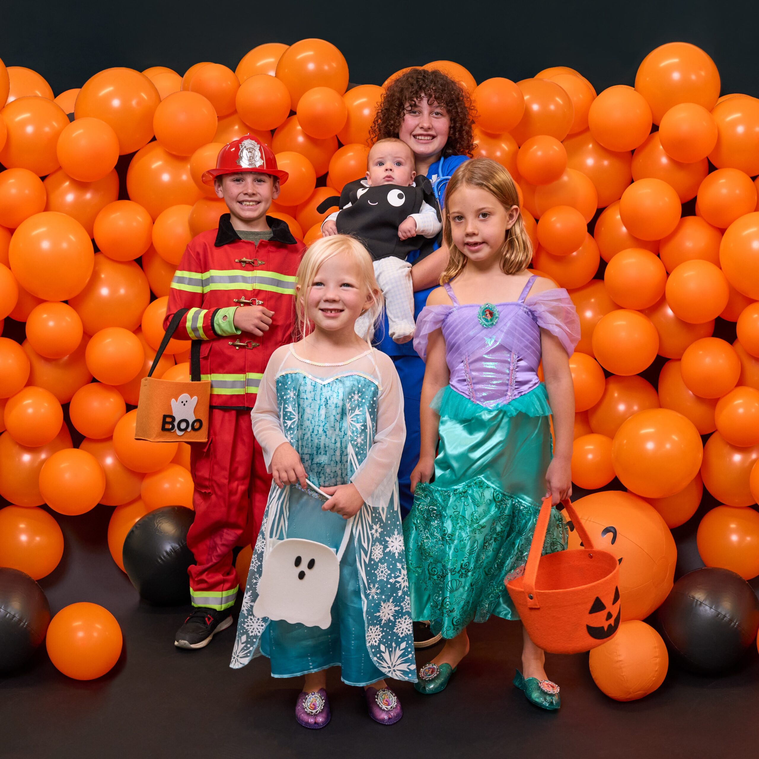 four children in halloween costumes standing in front of a pile of orange balloons against a black background